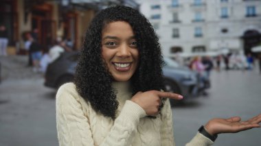 Kıvırcık saçlı, gülümseyen ve parmağıyla şehir caddesinde avucunu gösteren bir kadın. Joy Hope 'un keşfi merak uyandırıyor..