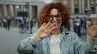 Dışarıdaki Vatikan ortamında gülümseyen bir kadın, arka planda insanlarla birlikte St. Peter Meydanı 'nda hareketli bir şehir atmosferi sergiliyor..