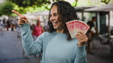 Restoran terasında gülümseyen bir kadın İsrail şekellerini elinde tutarken, bir yandan da paradan söz ediyor ve canlı bir ortamda paranın varlığını vurguluyor..