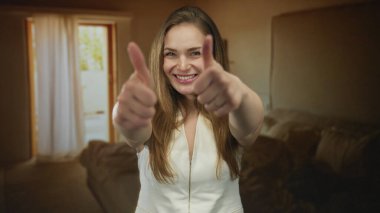 Beyazlar içinde beyaz elbiseli genç bir kadın aydınlık binada baş parmağını kaldırıyor ve göz kırparak gülümsüyor; iyimserlik.