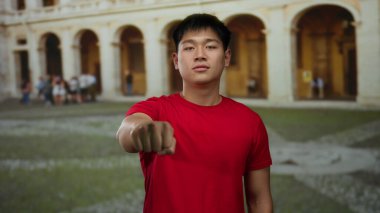 Kırmızı gömlekli genç adam, tarihi üniversitede, kemer yolları ve taş avlularla çevrili, azim ve kültürel gururu tasvir eden, açık havada kameraya yumruk uzattı..