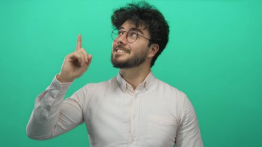 Sakallı genç adam yeşil bir arka plana karşı yukarıya doğru bakıyor, merak ve pozitiflik yayıyor..