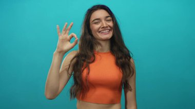 Turuncu bluz giyen bir kadın açık bir şekilde gülümserken deniz mavisi atölyesinde el işareti gösteriyor; iyimserlik..