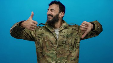 Ordu üniformalı İspanyol bir adam, sakallı, mavi arka planda bir aşağı bir yukarı hareket ediyor, modern asker hayatında karışık duygular ve ikiliği ifade ediyor..