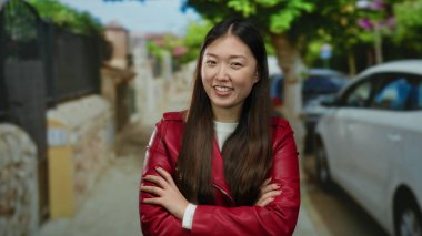 Kırmızı ceketli bir kadın yeşil ve park edilmiş arabalarla çevrili bir caddede gülümsüyor, canlı bir şehir atmosferini canlandırıyor..