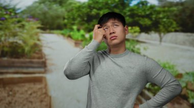 Parktaki genç adam gündüz vakti açık gökyüzünün altında yeşillik ve ağaçlarla çevrili gündelik kıyafetlerle düşünceli görünüyor..