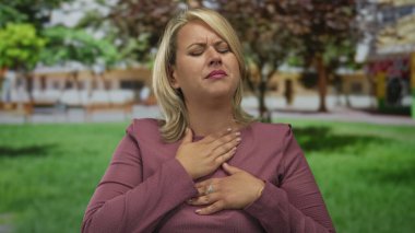 Pembe kazaklı genç sarışın kadın ağzını eliyle kapatıyor ve güneş ışığında yeşil parkta göğsünü tutuyor..