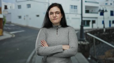 Esmer saçlı, gözlüklü ve gri kazaklı bir kadın açık alanda sakin bir ifadeyle şehir sokağında duruyor..