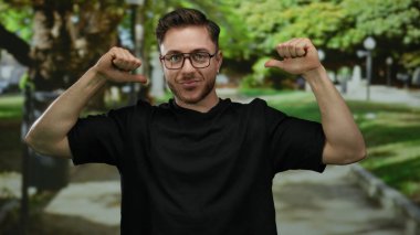 Parkta genç bir adam, siyah tişört giyiyor, başparmaklarıyla gülüyor ve kendini işaret ediyor, arka planda ağaçlar ve patikalar var, oyuncu, kendine güvenen bir tavır sergiliyor..