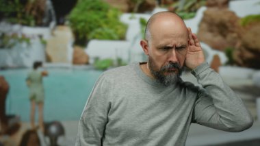 Sakallı ve kel kafalı bir adam açık havuz tesisinde dikkatlice dinliyor, sakin otel ortamını gösteriyor..