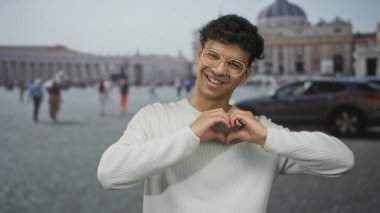 Beyaz süveter giyen ve gözlüklü bir adam Vatikan binasındaki kameraya işaret ediyor..