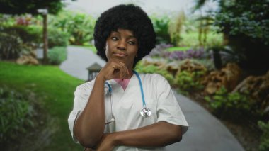 Genç Afro-Amerikan kadın doktor önlük ve steteskop giyerek yeşil parkta çene üstüne el, düşünceli bir yansıma..