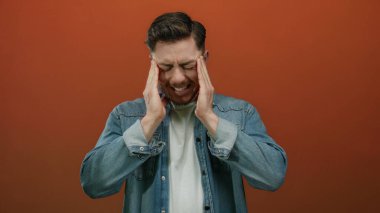 Hüsrana uğramış sakallı genç adam izole edilmiş kırmızı arka plan duvarına karşı rahatsızlık içinde başını tutuyor, baş ağrısı veya stresi ifade ediyor..