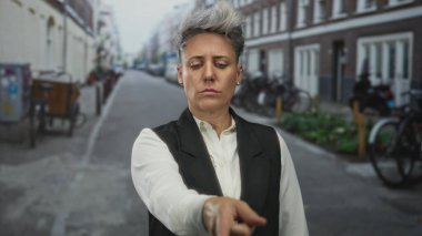 Kısa saçlı ve tarafsız ifadeli beyaz kadın, tuğla binalarla dolu bir caddeyi işaret ediyor. Kararlılık yönü:.