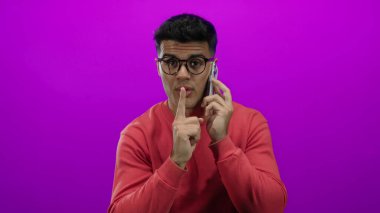 Kırmızı kazaklı yakışıklı adam telefonda canlı pembe arka planla ilgili konuşuyor. Sessizlik için jest yapıyor.