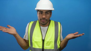 Çağdaş stüdyoda çelik yelek ve sert şapkalı bir adam avuçlarını mavi zemine yaslıyor; belirsizlik.