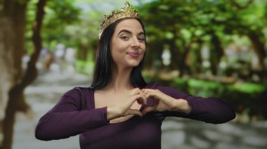 Parkta taç giyen bir kadın elleriyle kalp hareketi yapıyor, neşe ve kendini beğenmişlik gösteriyor, gür ağaçlarla çevrili, açık havada zarafet ve oyunculuğu somutlaştırıyor.