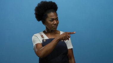 Önlük takan kadın mavi arka planda özgüvenli bir şekilde odak ve yönünü izole bir ortamda somutlaştırıyor, iddialı hareketlerle fikirleri işaret ediyor..