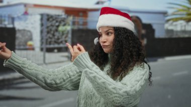 Şehir sokağında kazak giymiş, açık havada anlamlı tavırları ve neşeli tavırlarıyla samimi bir anı yakalayan Noel Baba şapkalı bir kadın..