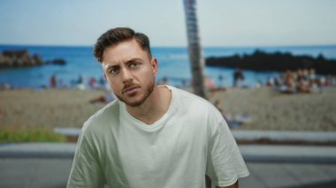 Sakallı genç adam arka planda deniz ve palmiye ağaçları olan bir sahilde duruyor, rahat bir deniz kenarı manzarası yakalıyor..