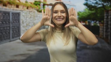 Mavi gökyüzünün altındaki şehir sokağında elleri çerçeveli gülümseyen kadın sıradan bir şehir ortamında mutluluk ve güven sergiliyor..