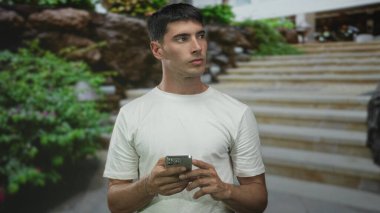 Beyaz gömlekli adam iki elinde akıllı telefon tutuyor ve yeşil yapraklı merdivenlere yan bakıyor; merak araştırma bağlantısı.