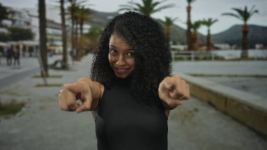 Siyah kıvırcık saçlı genç İspanyol kadın geniş bir gülümseme ve canlı caddede kamerayı işaret ediyor. Eğlenceli bir davet..