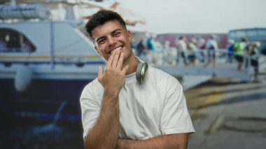 Sahilde bir teknenin yanında boynunda kulaklıklarla gülümseyen bir adam, arka planda insanlarla birlikte dışarıdaki bir ortamda samimiyet ve dostluk yayıyor..