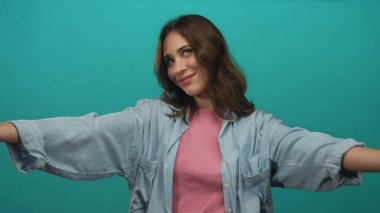 İzole edilmiş yeşil bir arka plana karşı kollarını açmış gülümseyen bir kadın günlük giysiler giyiyor. Portre tarzı bir ortamda dostluk ve mutluluk sergiliyor..