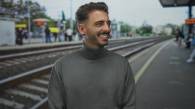 Trende gülümseyen ve bekleyen İspanyol genç bir adam tren istasyonunda gri balıkçı yakalı kazak giyiyor; memnuniyet.