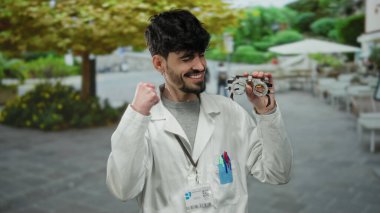 Açık havada gülümseyen beyaz önlüklü ve arka planda ağaçlar olan bir parkta gözlük takan genç bir adam, özgüven ve profesyonellik yayıyor..