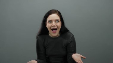 Esmer saçlı ve anlamlı yüzlü bir kadın, gri arka planda esmer uzun kollu bluz giyerken duygularını ve hareketlerini rahatça yansıtıyor..