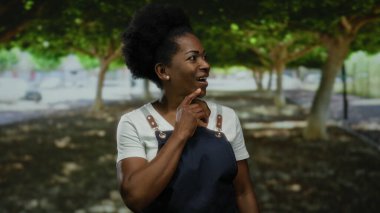 Önlük giymiş, gülümseyen ve yemyeşil bir parkta duran ağaçların arasından süzülen benekli güneş ışığıyla sıcak bir açık hava yaratan bir kadın..