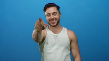 Sakallı, gülümseyen ve kameraya bakan genç bir adam izole edilmiş mavi bir arka plan duvarına bakıyor, kendine güveni ve nişanı yayıyor..