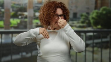 Şehir sokaklarında kötü kokuya tepki veren bir kadın, tiksindirici bir ifadeyle, gözlük takıyor, esmer saçlı, gün ortasında açık havada ortaya çıkıyor..