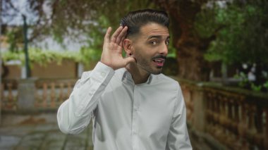 Beyaz gömlekli bir adam yeşil parkta bir patika boyunca bir taş balyaların yanında dururken kulağını kapar; merak.