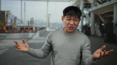 Şehir sokaklarında duygusal olarak el kol hareketi yapan genç Çinli bir adam, gri bir kazak giyiyor, arka planda modern şehir mimarisi var..