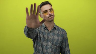 Güneş gözlüklü genç adam, parlak sarı arkaplan karşısında el işaretini uzatarak, izole bir ortamda uyarı veya reddetme mesajı iletiyor..