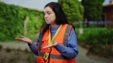 Turuncu yelekli bir kadın dışarıda yeşil bir park ortamında düşünceli bir ifadeyle duruyor. Toplum hizmetine bağlılığını ve çevreye bağlılığını gösteriyor..