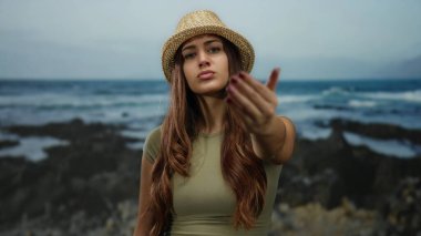 Deniz kıyısında jest yapmaya davet eden kadın, sakin okyanus manzarasına tepeden bakan ciddi bir ifadeyle hasır şapka takıyor.
