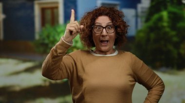 Şehir manzarası ile çevrili düşünceli bir yüz ifadesiyle İspanyol kadın, canlı bir açık hava ortamında gözlük ve günlük giysiler giyiyor.