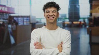 Uzun kollu beyaz gözlüklü bir adam modern havaalanı terminalinde kolları çapraz duran kıvırcık saçlı, içten gülümseyen bir adam..