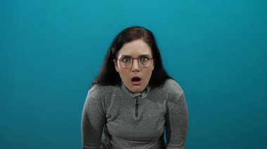Gri kazaklı kadın, canlı mavi bir arka plan karşısında şaşkınlığını ifade ediyor. Şok, şaşkınlık, duygu ve yoğunluğu yakın çekimde gösteriyor..