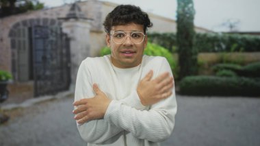 Genç adam, hafif süveter içinde titrerken taş bina girişinin dışında çıplak omuzlarına sarılıyor..