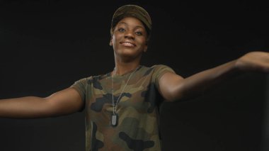 Kamuflaj şapkası takan ve künyeli bir kadın stüdyo ortamında göğüs hizasında iki elini de uzatıyor..