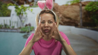 Pembe gömlek ve tavşan kulaklığı takan kadın, gün ışığında binanın önünde havuz başında elleri olan ağız çerçevesi takıyor..