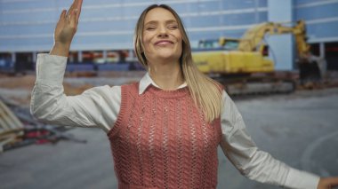 Dışarıdaki inşaat alanında kazıcıyla gülümseyen, kaya jestleri yapan, pembe kazak giyen, bina ortamında heyecan ve neşeyi simgeleyen bir kadın..