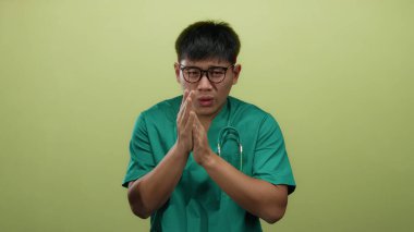 Önlüklü genç adam, gözlüklü ve steteskoplu izole edilmiş sarı arka plana karşı üşümüş hissediyor. Elleri sımsıkı bağlı, soğuk bir ortamda rahatsızlık gösteriyor.