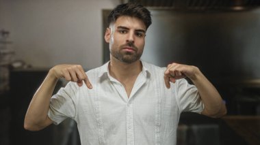 Sakallı ve odaklanmış bakışlı bir binanın içindeki beyaz gömleğini aşağı doğru gösteren genç İspanyol adam..