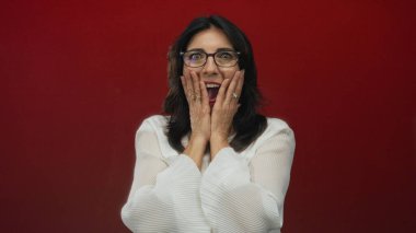 Elleri yanaklarında ve kırmızı stüdyoda ağzı açık, gözlüklü ve beyaz bluzlu bir kadın. Sürpriz..